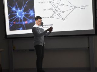 Prelegent gestykuluje na tle ilustracji neuronów i schematu sieci.