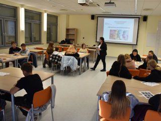 Prowadząca warsztaty stoi między grupami uczniów przy połączonych ławkach.