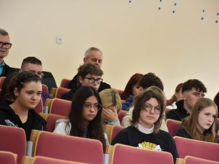 Uczestnicy wykładu siedzą w rzędach czerwonych foteli w auli.