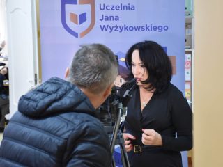 Kobieta rozmawia z operatorem kamery przed banerem Uczelni Jana Wyżykowskiego.