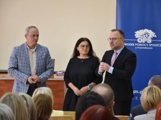 Mężczyzna w garniturze przemawia do mikrofonu obok kobiety i mężczyzny w marynarce w kratę.
