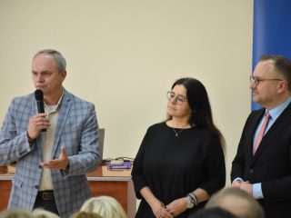 Mówca w marynarce w kratę gestykuluje podczas wystąpienia przed publicznością.