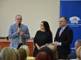 Mężczyzna w marynarce w kratę przemawia obok dwojga przedstawicieli organizatorów.