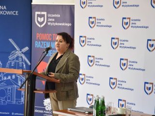 Prelegentka w marynarce w kratę przemawia przy banerach Gminy Polkowice i UJW.