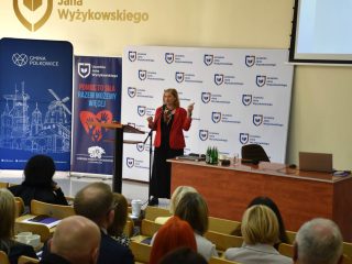 Prelegentka w czerwonej marynarce gestykuluje podczas wystąpienia przed publicznością.