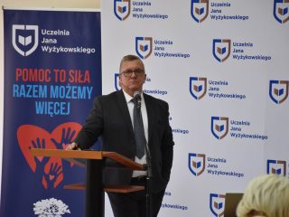 Mężczyzna w okularach przemawia na tle banera „Pomoc to siła, razem możemy więcej”.