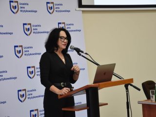 Prelegentka w czarnej sukience przemawia przy mównicy z laptopem.