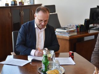 Mężczyzna w granatowej marynarce podpisuje dokument przy biurowym stole.