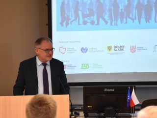 Mężczyzna w okularach prowadzi wystąpienie na tle ekranu z patronami konferencji.