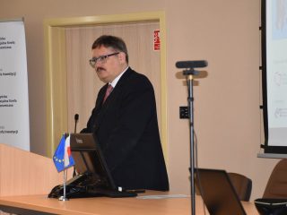 Mężczyzna z wąsami przemawia przy mównicy z flagami Polski i Unii Europejskiej.