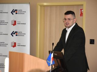Prelegent w okularach stoi przy mównicy obok banera Legnickiej Specjalnej Strefy Ekonomicznej.
