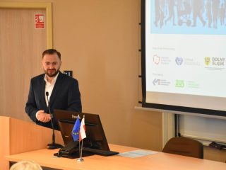 Brodaty mężczyzna przemawia przy mównicy na tle slajdu konferencji.