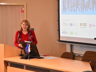 Prelegentka w czerwonej bluzce przemawia przy mównicy podczas konferencji.