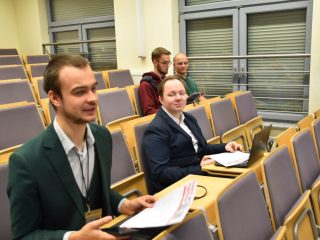 Uczestnicy konferencji siedzą w sali wykładowej z dokumentami i laptopem.