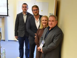 Czworo uczestników konferencji dotyczącej zarządzania danymi pozuje w sali wykładowej.