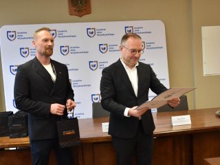 Mężczyzna w okularach odczytuje certyfikat; obok stoi mężczyzna z torbą upominkową.