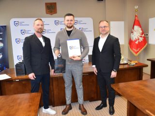 Wyróżniony w szarym swetrze prezentuje certyfikat i torbę upominkową na wspólnym zdjęciu.