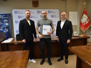 Mężczyzna w szarej koszulce prezentuje certyfikat pomiędzy dwoma przedstawicielami.