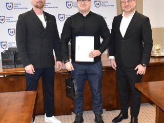 Wyróżniony w okularach prezentuje certyfikat na wspólnym zdjęciu z wręczającymi.