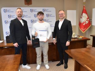 Wyróżniony w białej koszulce prezentuje certyfikat i torbę upominkową.