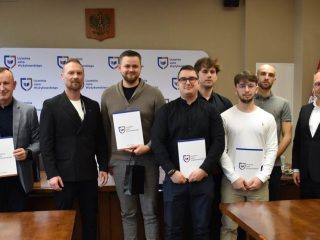 Grupa uczestników uroczystości KGHM pozuje z certyfikatami przed ścianką UJW.