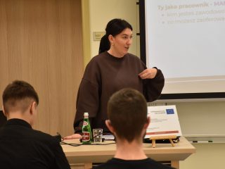 Prowadząca warsztaty dotyczące CV przemawia przy ekranie do uczestników siedzących przy stołach.