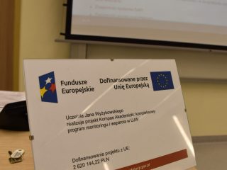 Tabliczka o dofinansowaniu projektu UJW z Funduszy Europejskich na stole w sali warsztatowej.