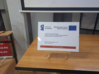 Tabliczka informująca o dofinansowaniu projektu UJW z Funduszy Europejskich, ustawiona na stoliku.