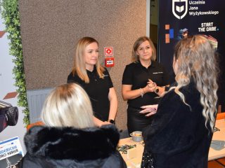Przedstawicielki pracodawcy słuchają uczestniczki spotkania przy stole informacyjnym.