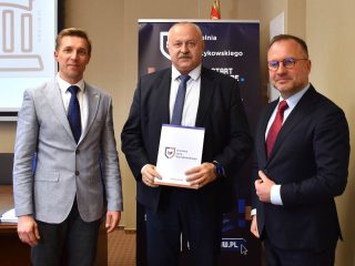 Mężczyzna z wąsami prezentuje dokument z logo UJW, stojąc pomiędzy dwoma przedstawicielami uczelni.
