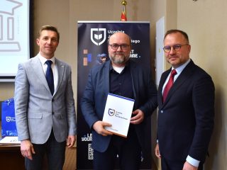 Brodaty mężczyzna w okularach pozuje z dokumentem z logo UJW pomiędzy dwoma mężczyznami.