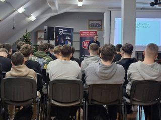 Uczniowie siedzą w rzędach przed ekranem i banerami UJW oraz szkoły.