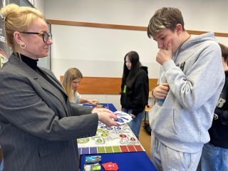 Prowadząca wyjaśnia zadanie uczniowi w szarej bluzie przy planszy gry.