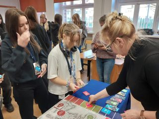 Dwie uczennice obserwują prowadzącą wskazującą pola na planszy.
