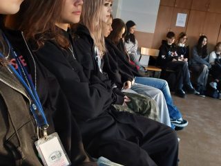 Uczestnicy warsztatów z identyfikatorami siedzą w rzędzie wzdłuż ściany.