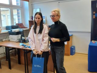Prowadząca i uczestniczka z niebieską torbą upominkową pozują w sali.