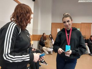 Dwie uczestniczki gry rozmawiają, trzymając karty i kolorowe identyfikatory.