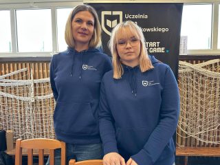 Dwie przedstawicielki uczelni w granatowych bluzach z logo UJW przy stoisku targowym.