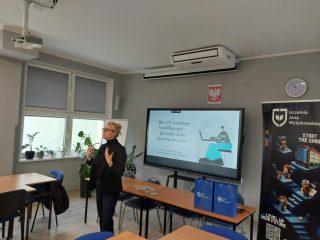 Prowadząca prezentację dla uczniów stoi przy ekranie i banerze UJW w sali lekcyjnej.