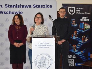 Kobieta przemawia do mikrofonu przy mównicy I Zespołu Szkół we Wschowie, w towarzystwie dwóch osób.