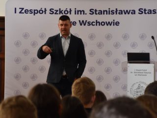 Prelegent gestykuluje przed publicznością na tle ścianki szkoły we Wschowie.
