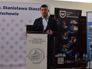 Prelegent przy laptopie na mównicy; nad nim slajd z zielonym schematem.