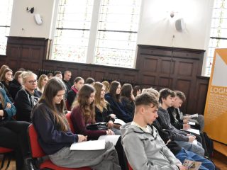 Uczniowie siedzą w rzędach przy witrażowych oknach auli podczas Dnia Logistyka.