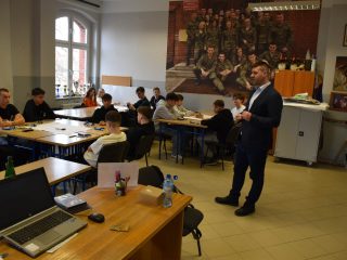 Prowadzący warsztaty stoi między stolikami z pracującymi grupami uczniów.