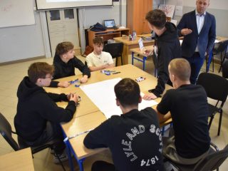 Uczniowie omawiają zadanie przy wspólnym arkuszu, obok stoi prowadzący warsztaty.