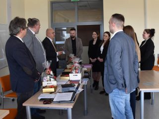 Rozmowa uczestników obron z komisją przy stole z dokumentami i upominkami.