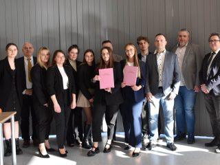 Grupa uczestników obron inżynierskich pozuje w sali; dwie osoby trzymają różowe prace dyplomowe.