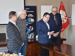 Czterej członkowie komisji stoją przy stole; mężczyzna w granatowym garniturze odczytuje dokument.