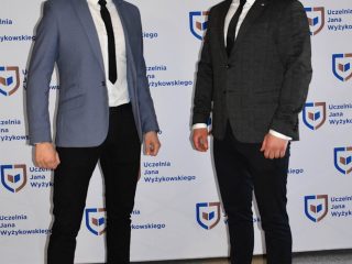 Dwaj mężczyźni w garniturach pozują na tle ścianki z logo UJW podczas obron inżynierskich.