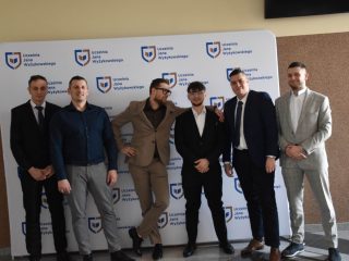Sześcioosobowa grupa absolwentów podczas wspólnego zdjęcia na tle logo UJW.